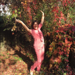 Auf diesem Bild wirft eine in rot gekleidete Frau, im Herbstlaub stehend, voller Freude die Arme hoch mit einem ansteckendem Lächeln im Gesicht.